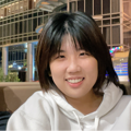Young woman with short black hair, wearing a white hoodie, smiling at the camera in a cozy indoor cafe setting.